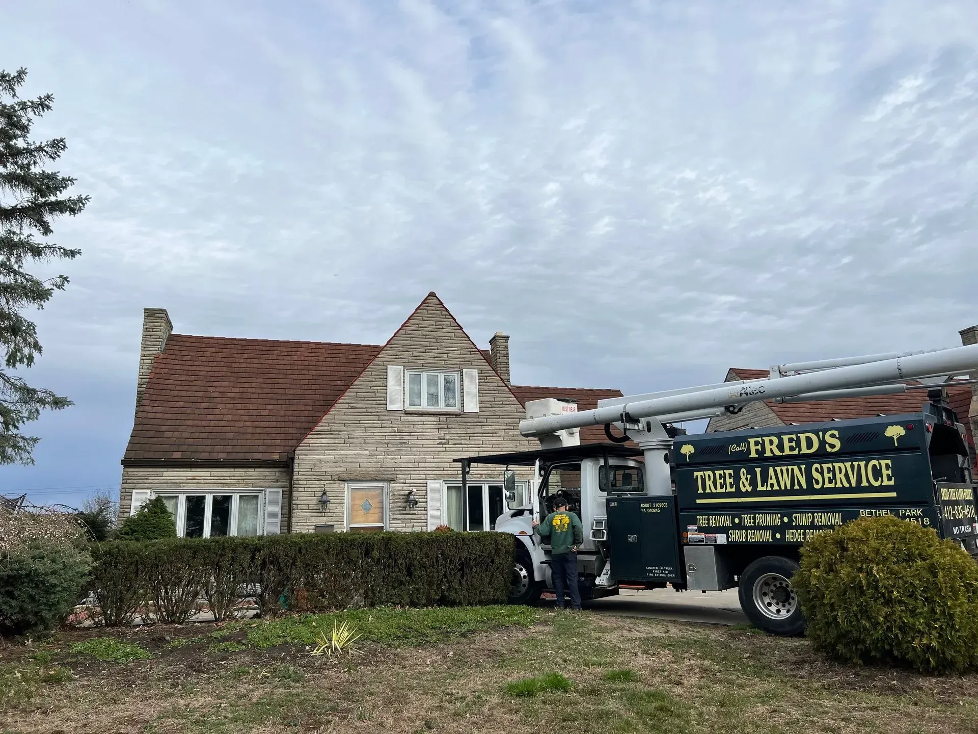 A landscaping truck parked in front of a stone house. Person standing beside the truck. Overcast sky.