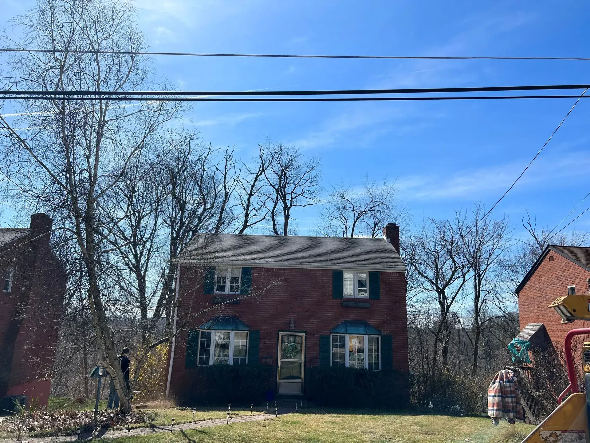 Red brick house with green shutters and a front door, trees, and power lines against a blue sky.