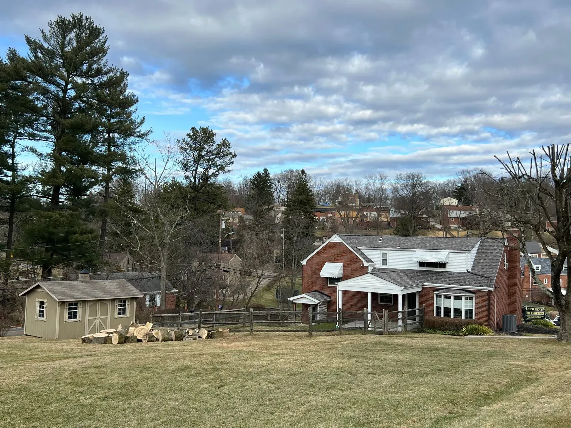 Brick house with porch, small shed, fenced yard, and sheep under a cloudy sky.
