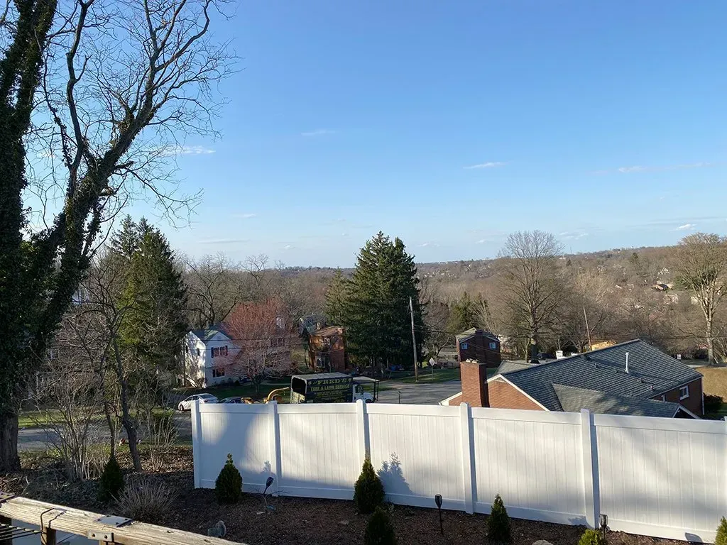 White fence in front of a residential area with trees under a blue sky.