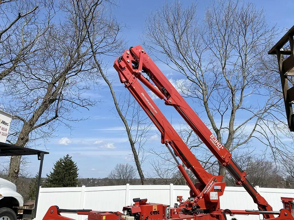 Red boom lift extended against a blue sky, trees, and a white fence.
