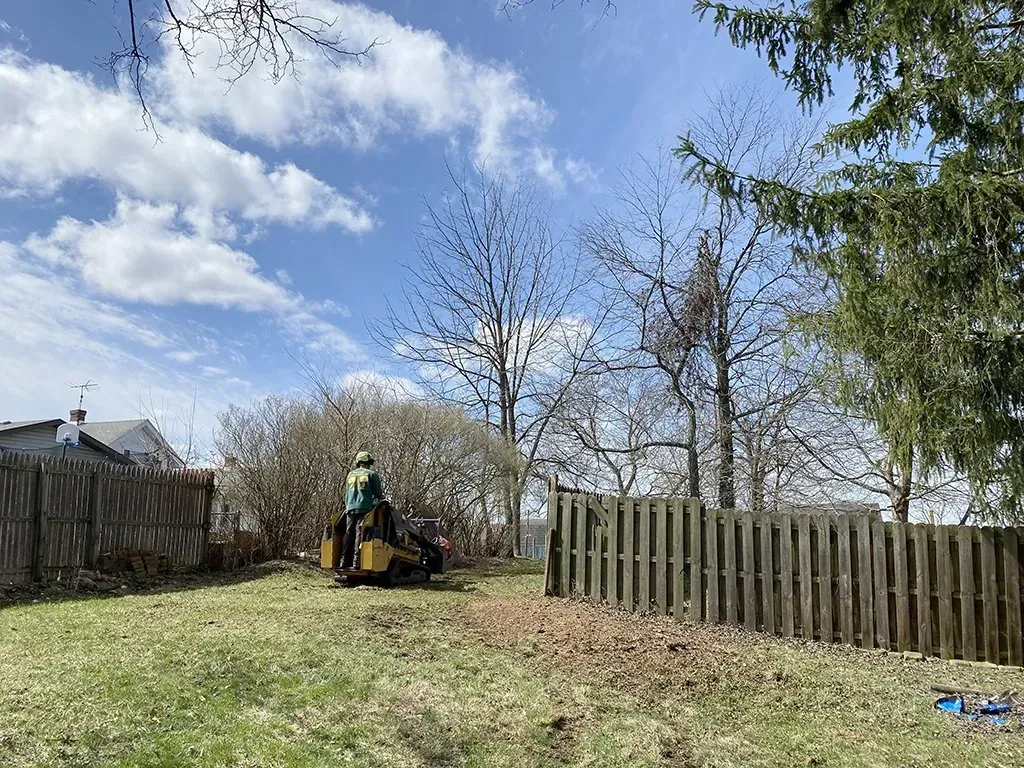 Person operating a wood chipper in a grassy backyard near a wooden fence on a sunny day.