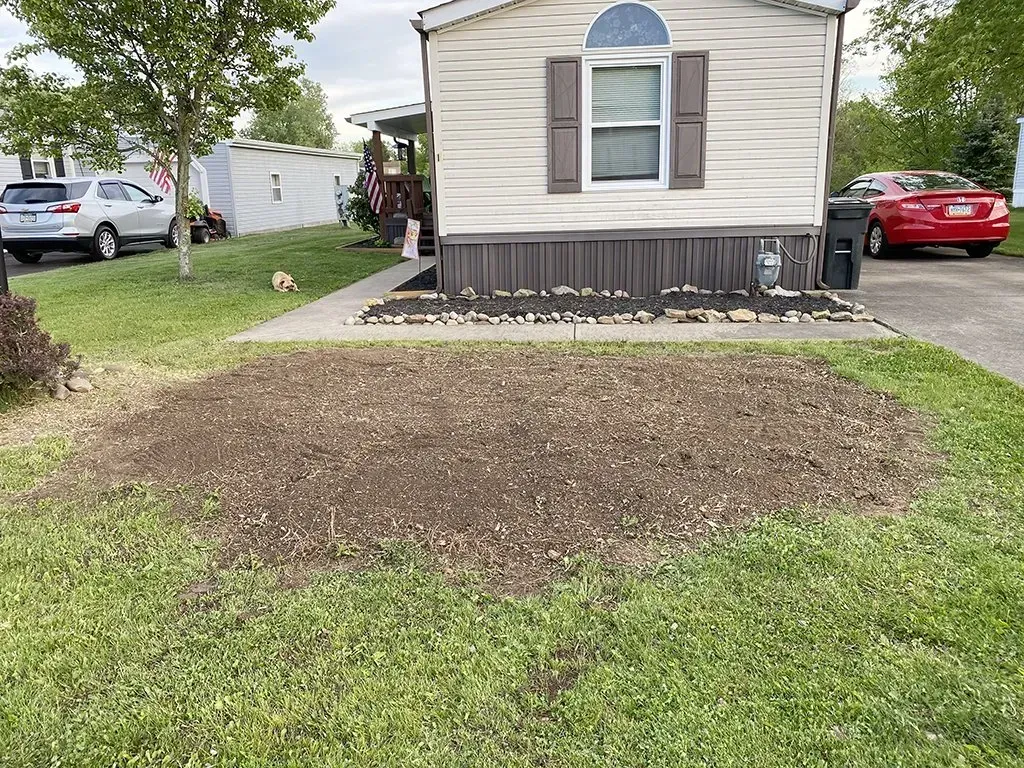 A mobile home with a dirt patch in front, surrounded by grass and a driveway.