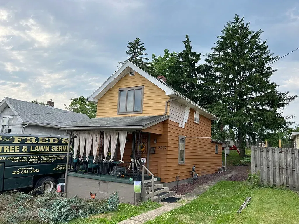 Small yellow and brown house with porch, next to a tree and a truck.