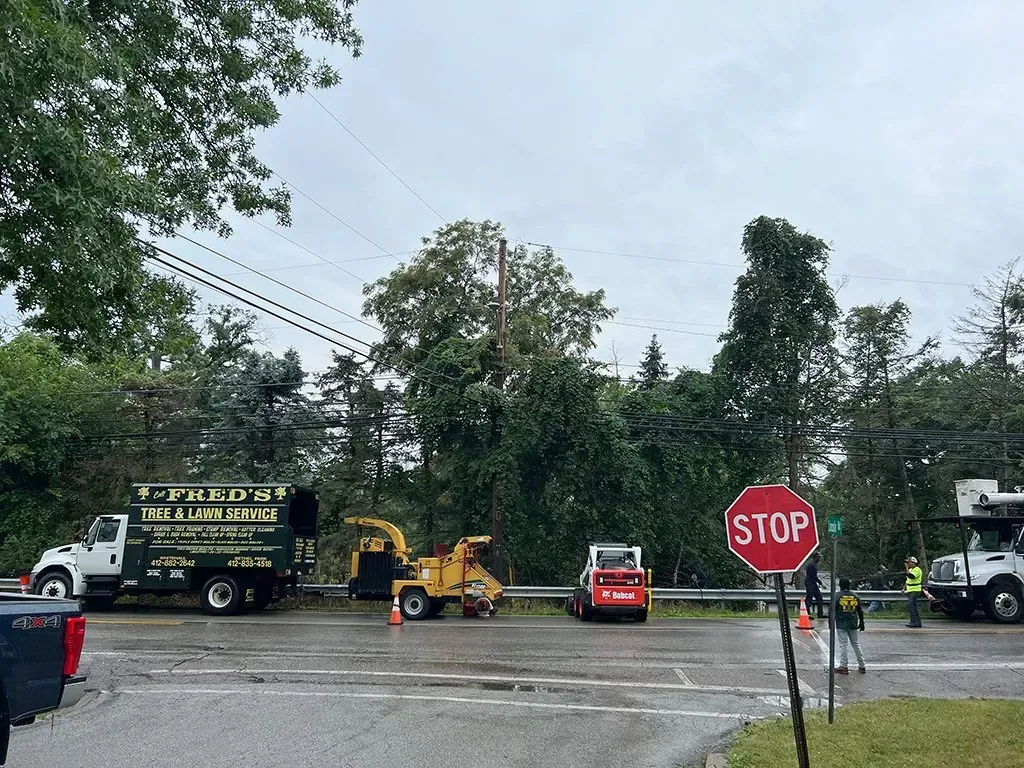 Crews working on power lines with trucks, a chipper, and a skid steer at an intersection under cloudy skies.