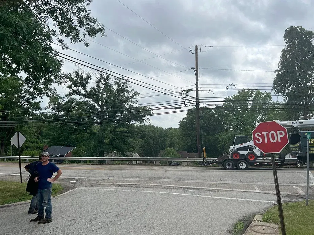 Man beside a road with a stop sign; a tractor sits on a trailer. Overcast day.