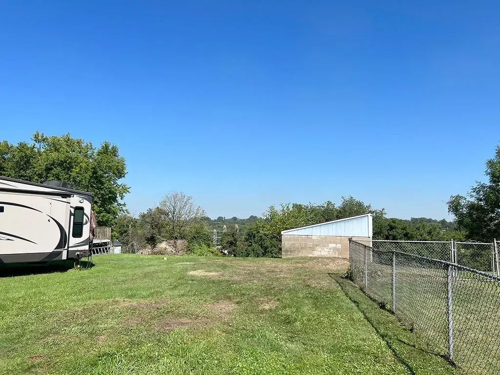 Grassy yard with a trailer on the left, chain link fence on the right, and a building in the distance under a clear blue sky.