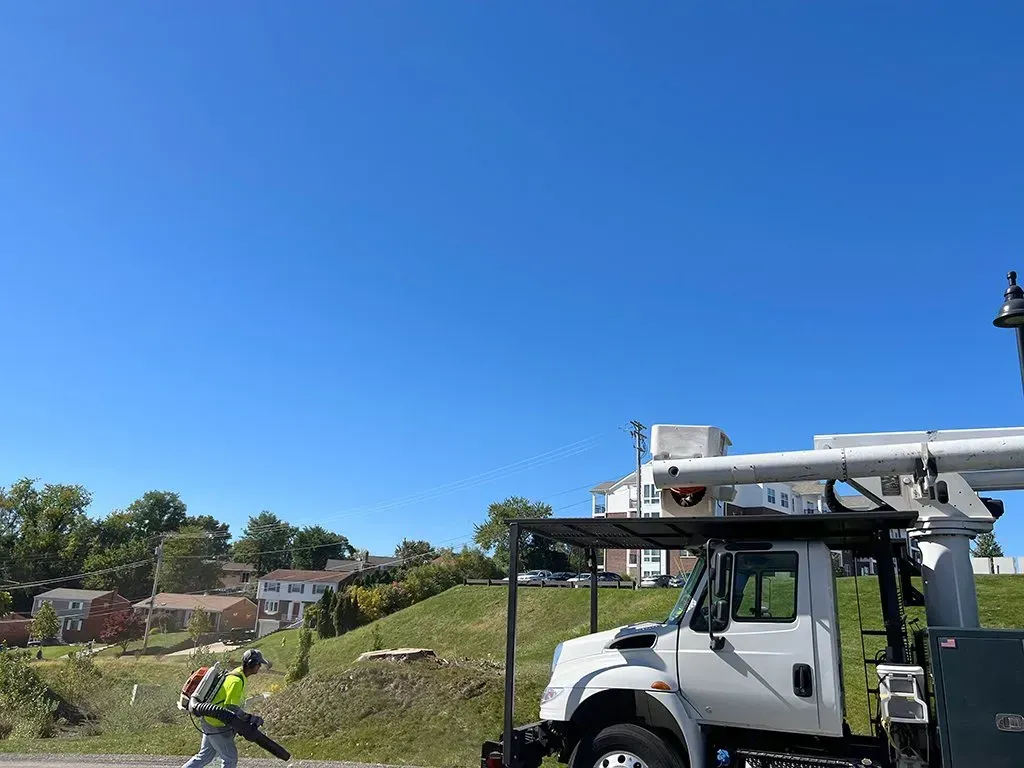 A worker blowing leaves with a backpack blower near a white utility truck on a sunny day. Houses in background.