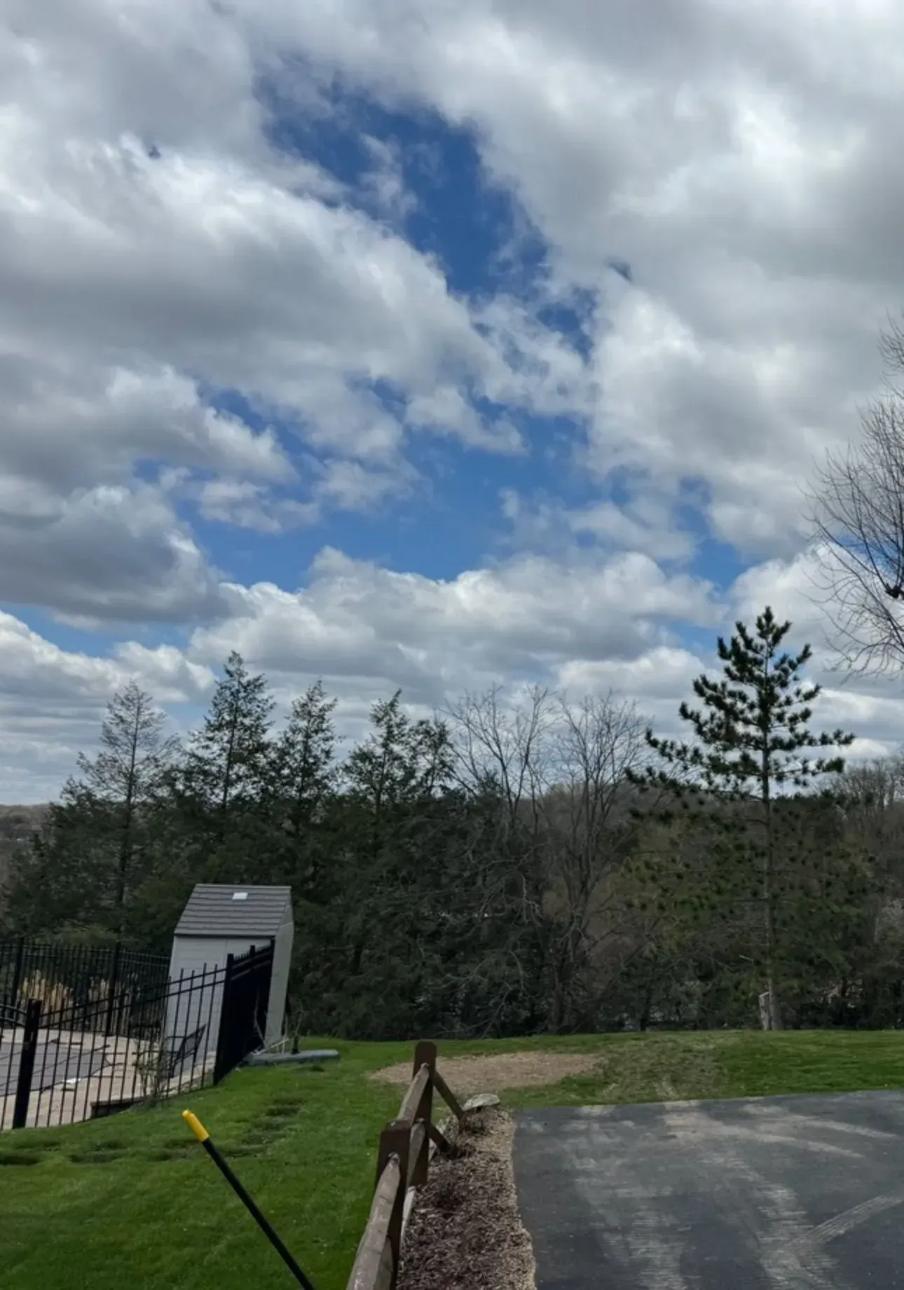 Blue sky with scattered clouds over trees, a small shed, and a grassy area.