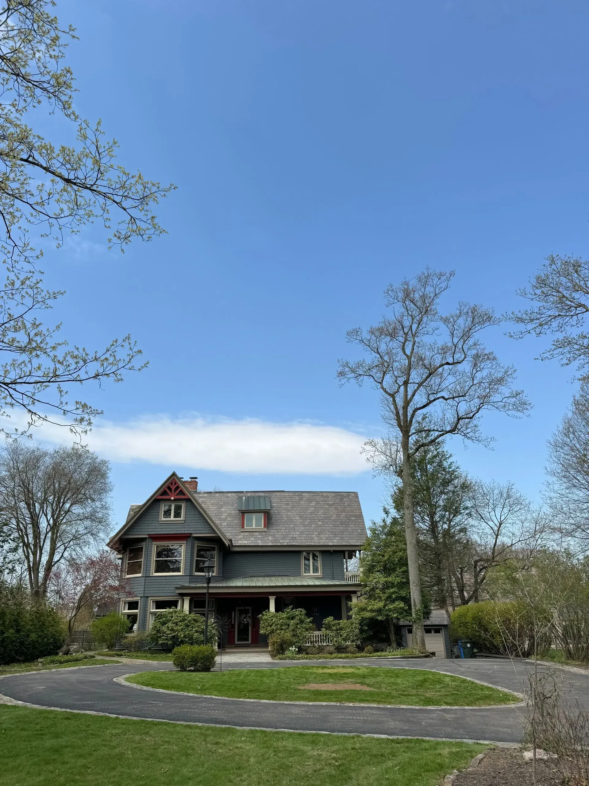 Two-story grey house with a dark red roof and trim, on a circular driveway, under a bright blue sky.