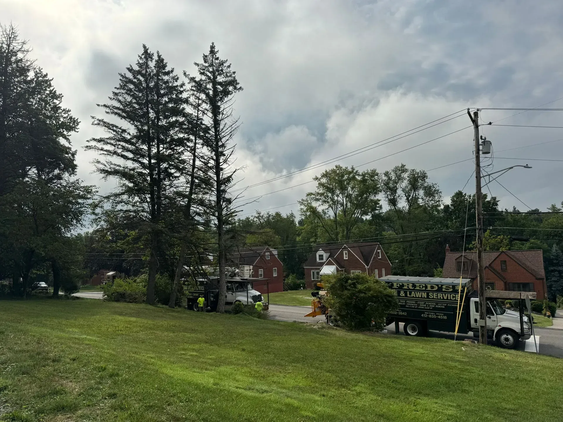 A utility truck parked near houses, trees, and power lines, on a grassy hill under a cloudy sky.
