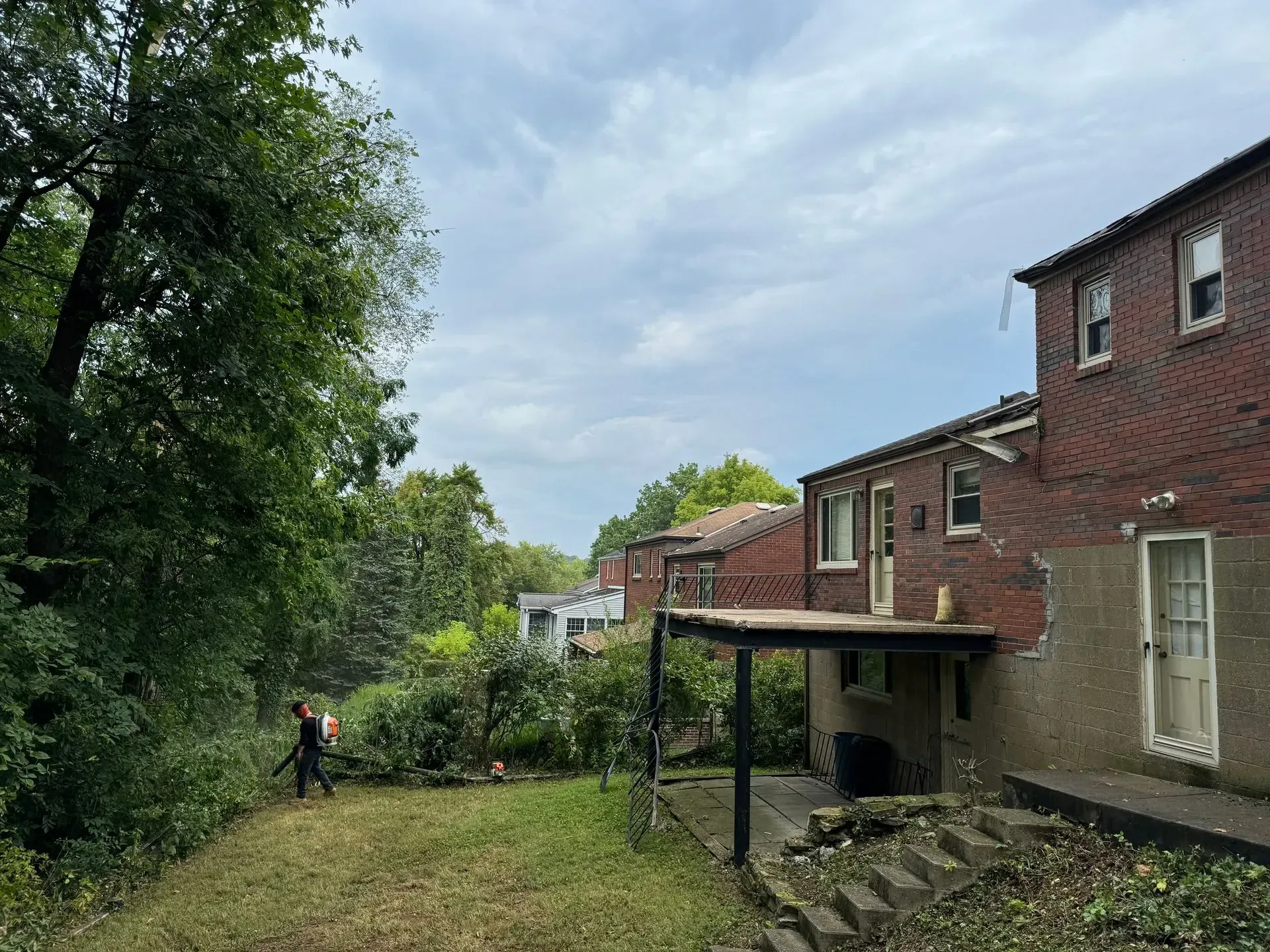 Person trimming overgrown yard next to red brick buildings under a cloudy sky.