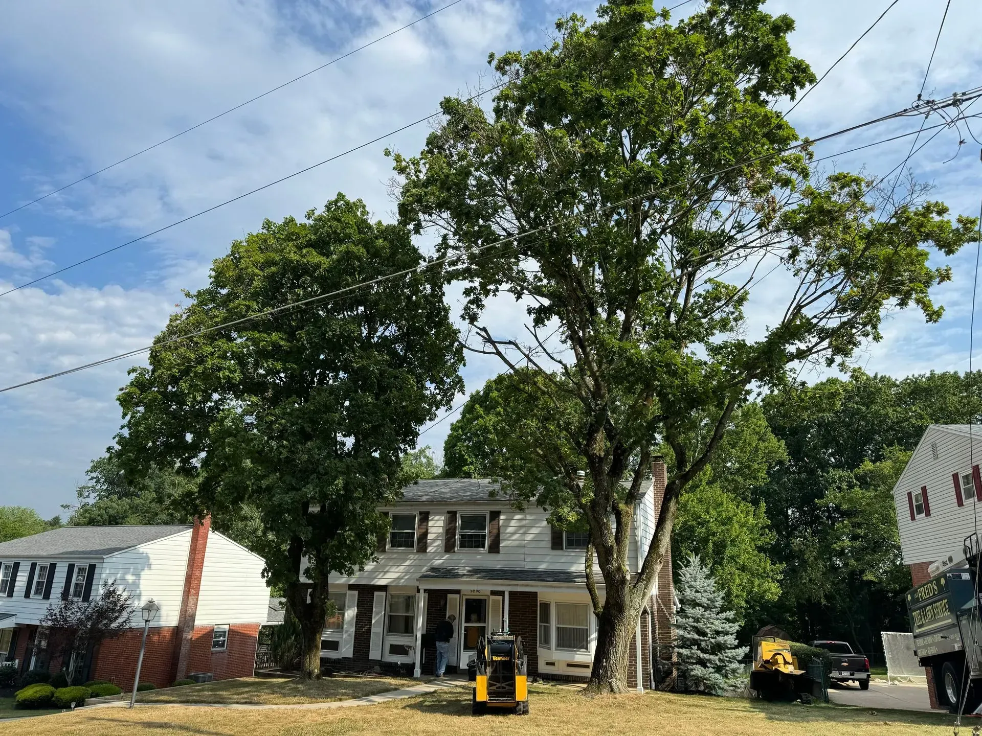 Two-story house with trees; a small construction vehicle in front. Blue sky and power lines overhead.