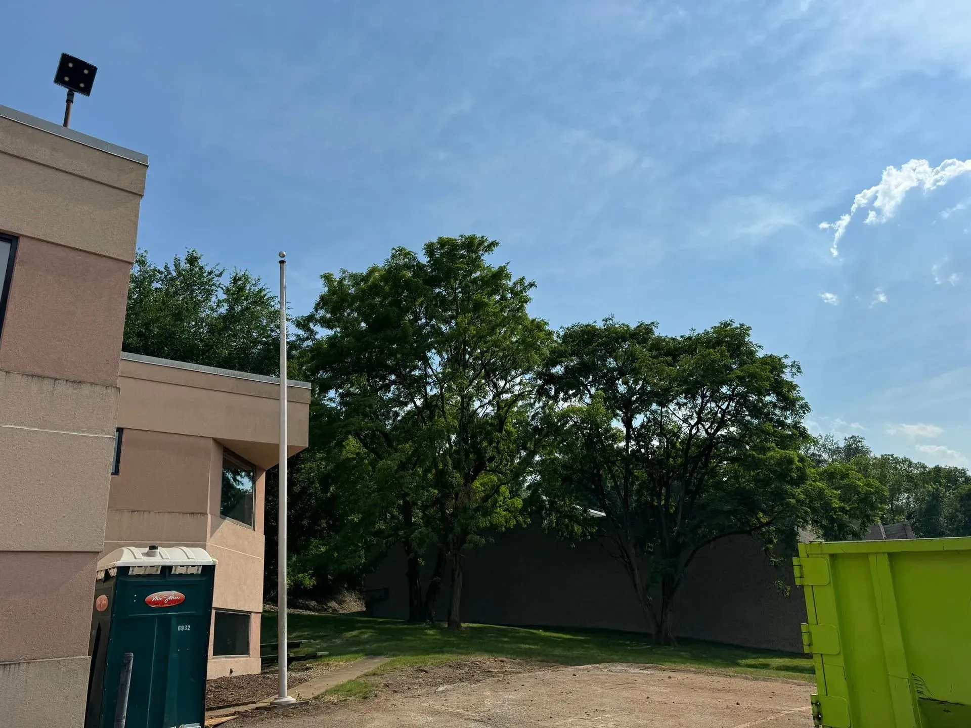 Exterior of building with flagpole, trees, and blue sky. A portable toilet and dumpster are visible.