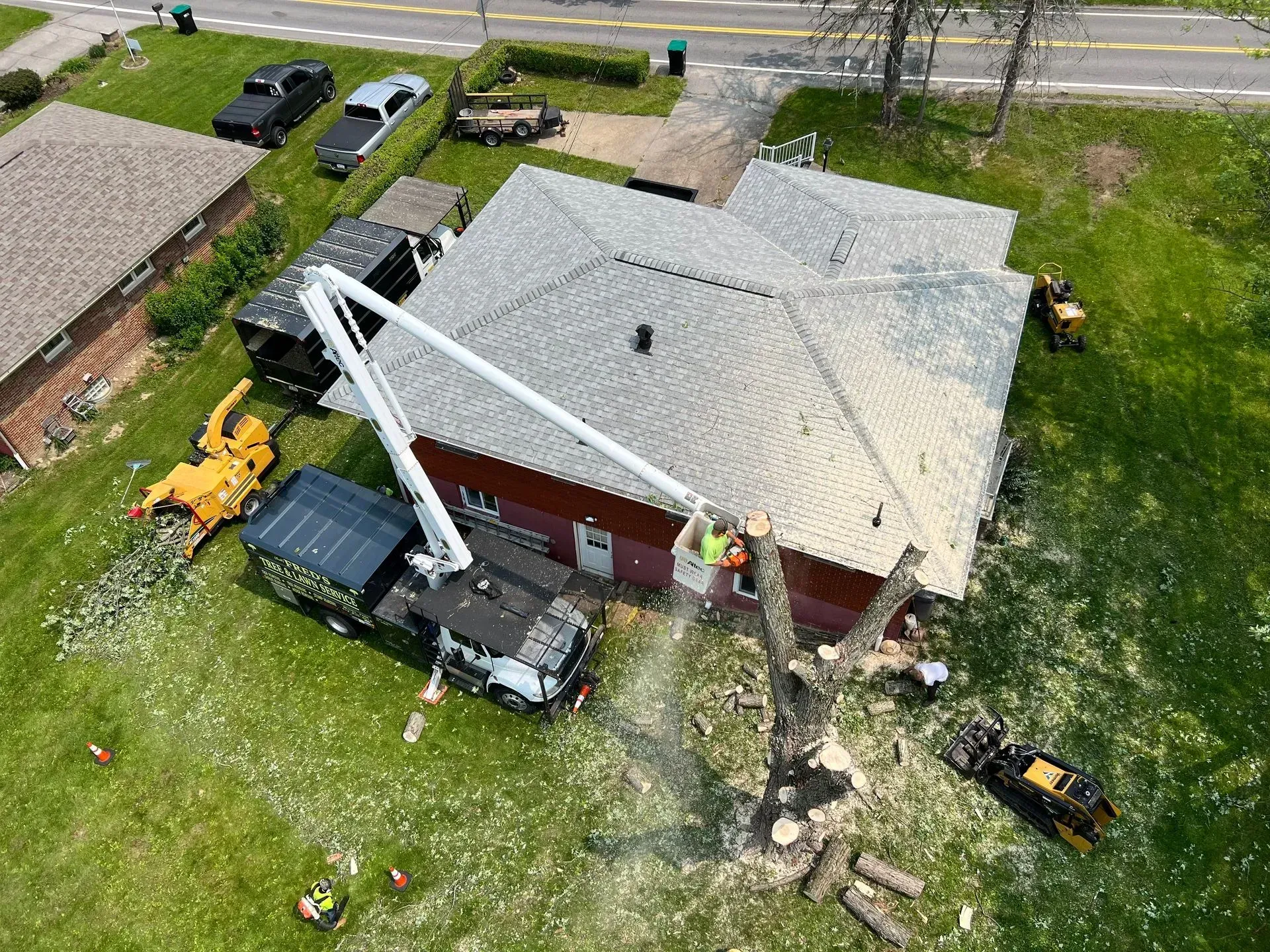 Tree removal in progress: workers in a lift trim branches near a red house. Green lawn, truck, and equipment visible.
