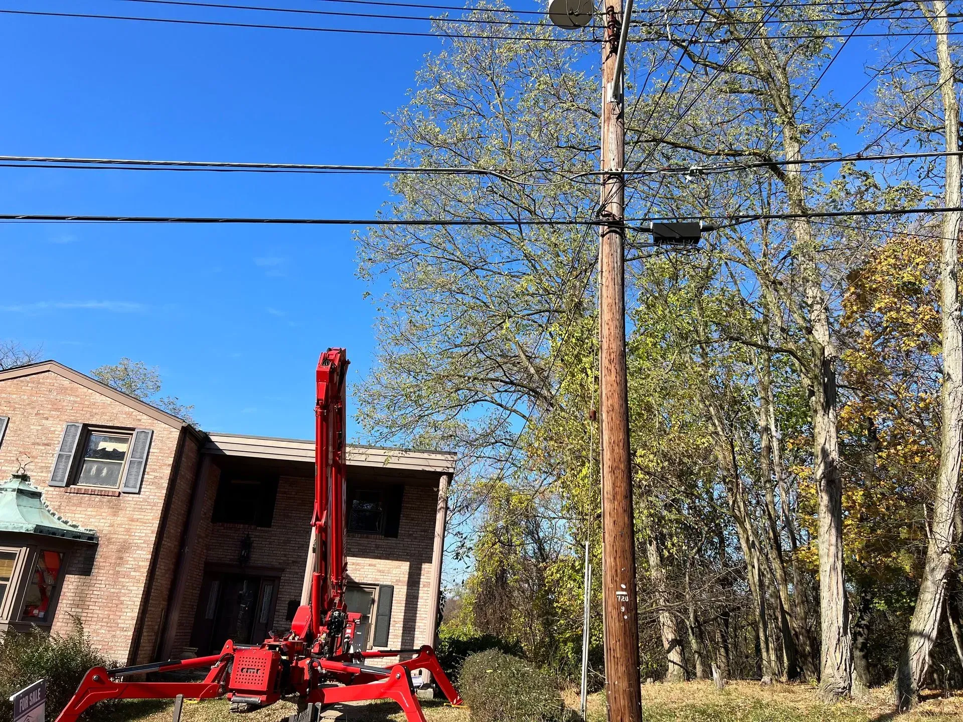 A red crane near a utility pole with power lines, trees, and a house under a blue sky.