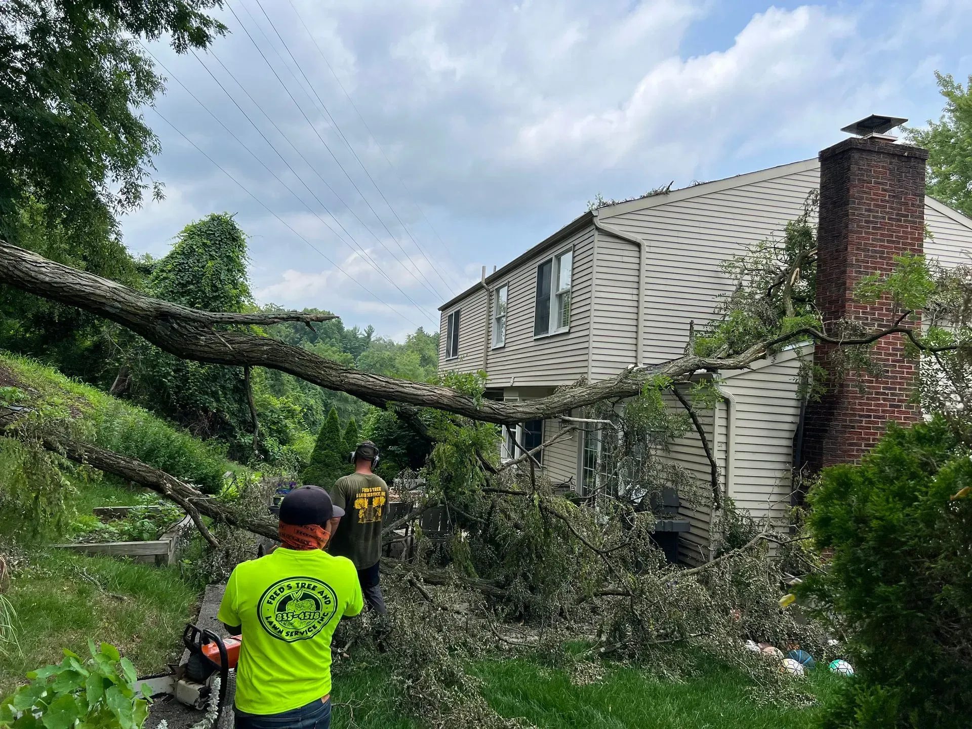 Men cut a fallen tree off a house with a chainsaw. Green grass, cloudy sky, brick chimney.