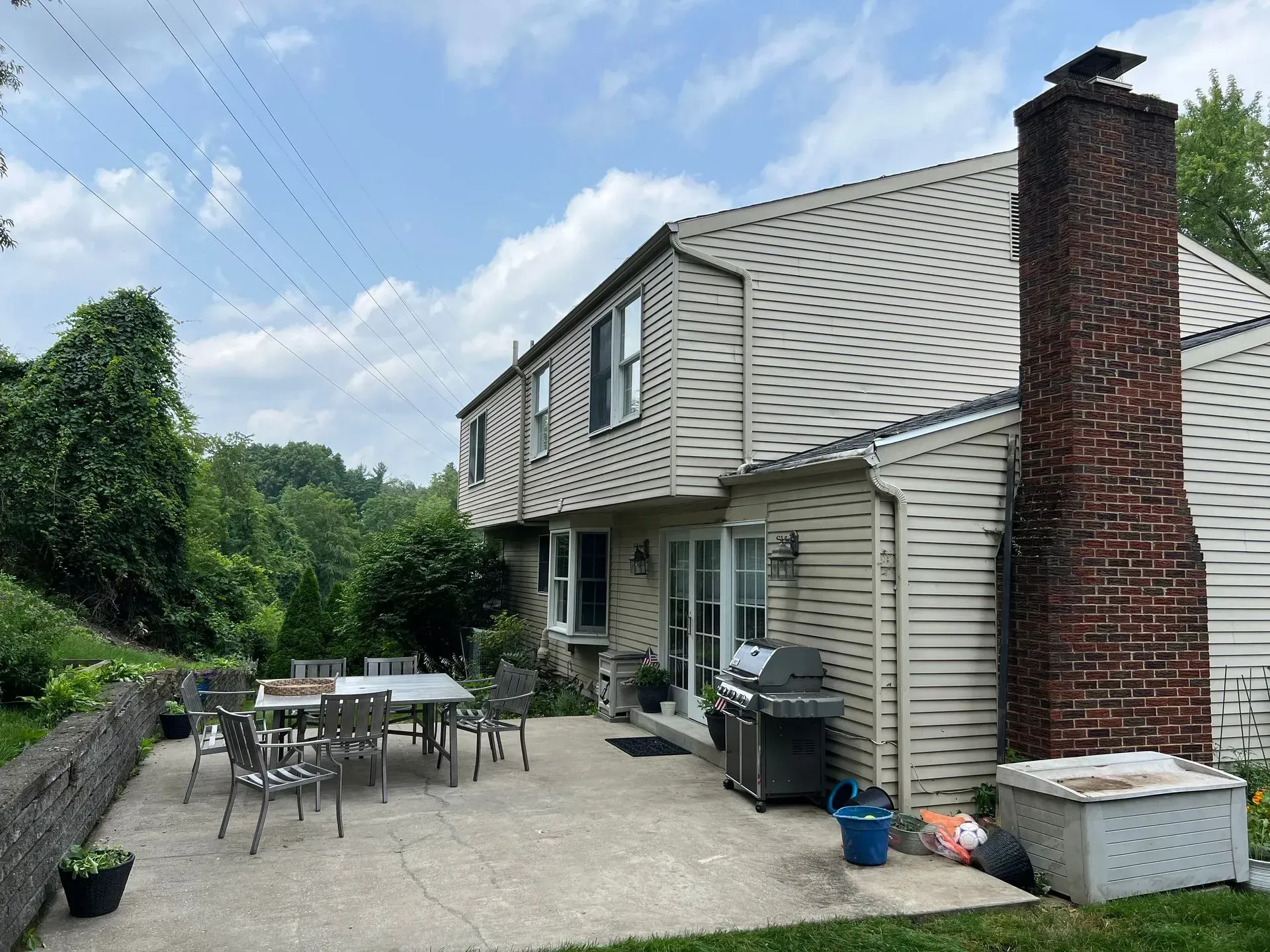 Backyard patio with table, chairs, grill, and two-story house with brick chimney and greenery.
