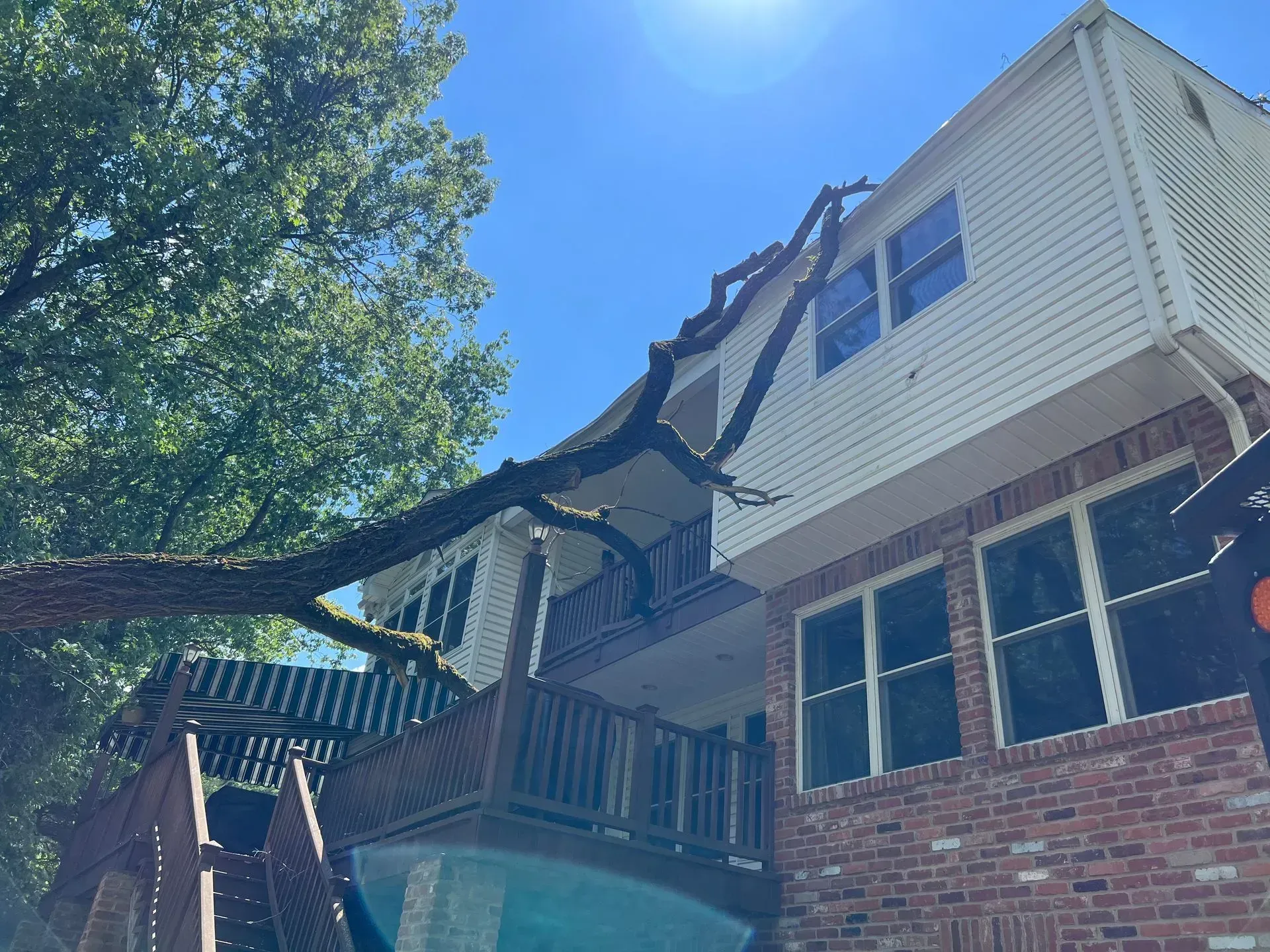 Tree branch fallen on a two-story house with a deck. Blue sky overhead.