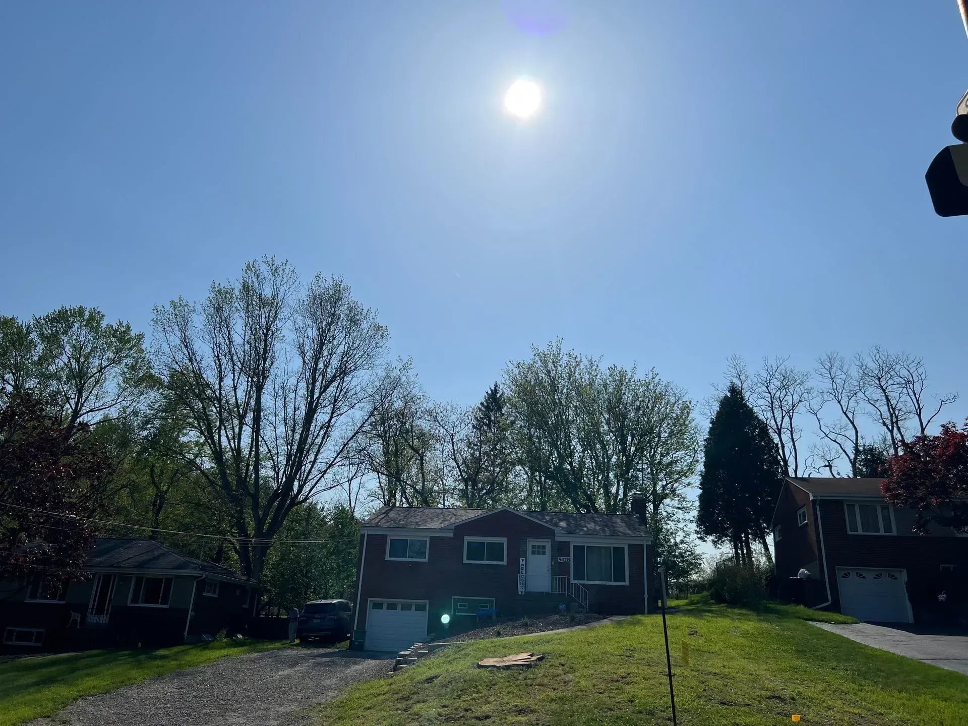 Suburban houses under a bright blue sky with a sun shining overhead, trees and greenery.