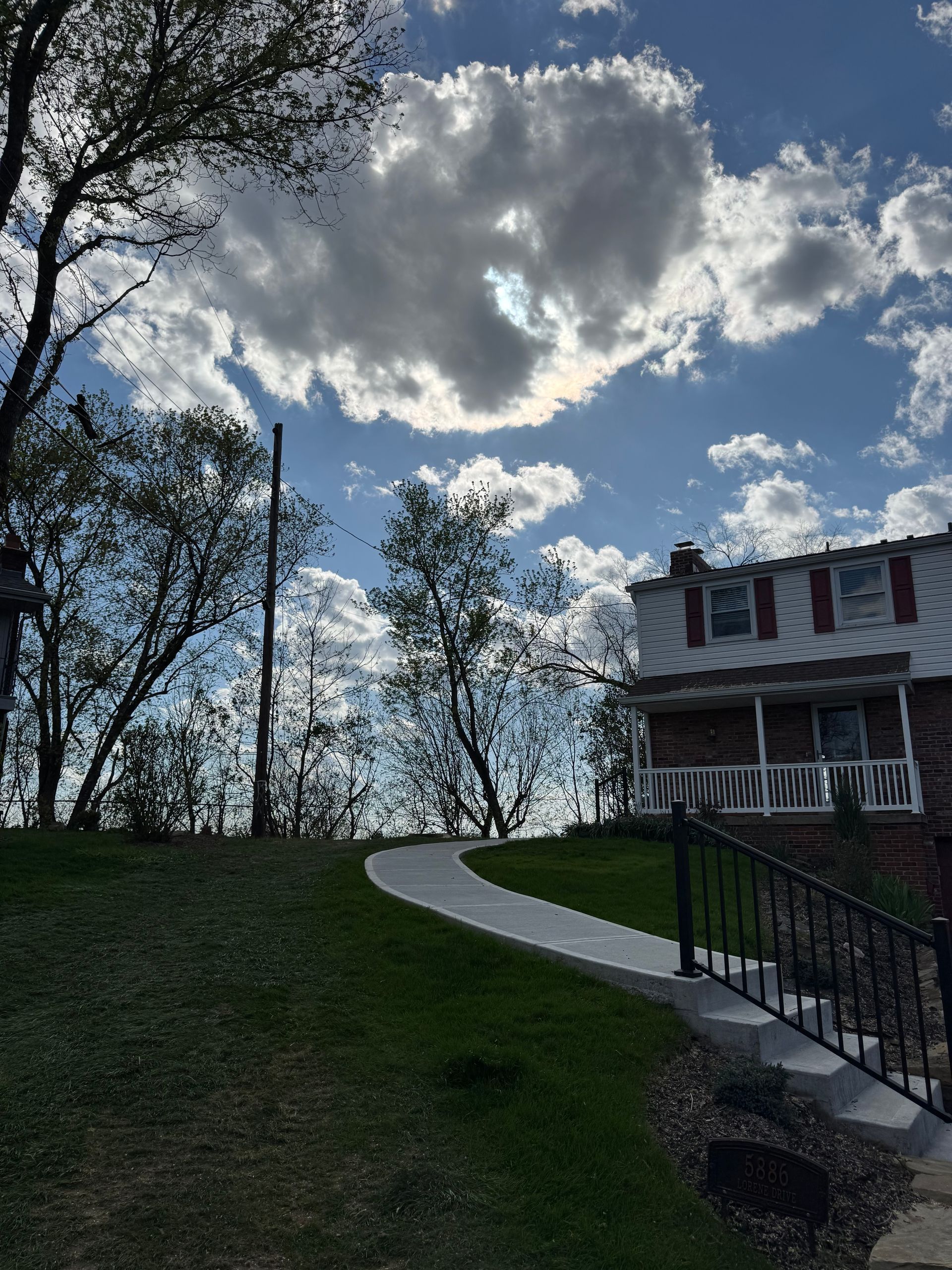 Curved walkway up a grassy hill beside a house, with trees silhouetted against a cloudy sky.