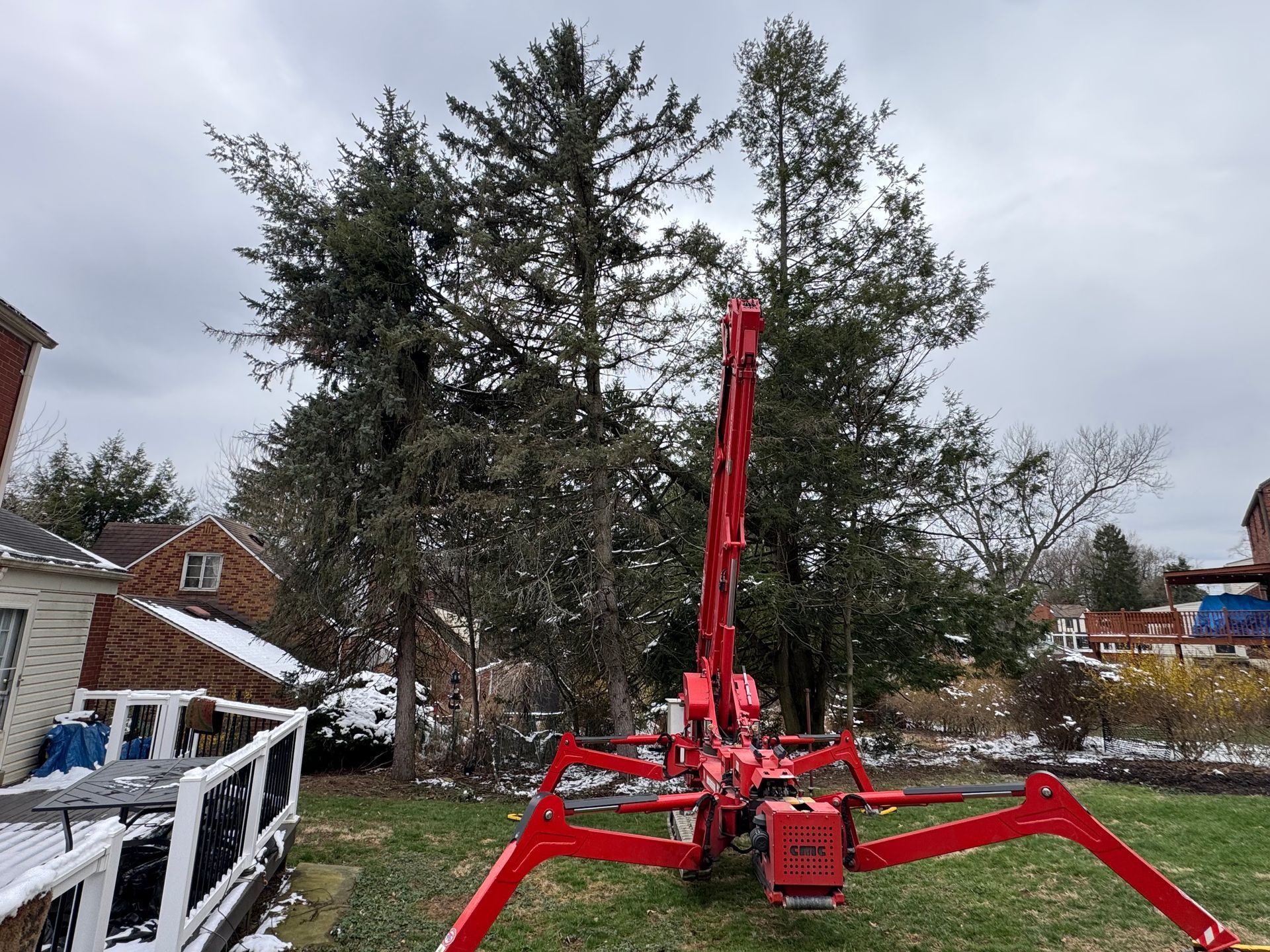 Red forestry machine with extended arms in a yard beside trees and houses under a cloudy sky