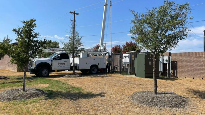 White utility truck parked beside a brick wall and utility poles in a dry yard under a clear blue sky