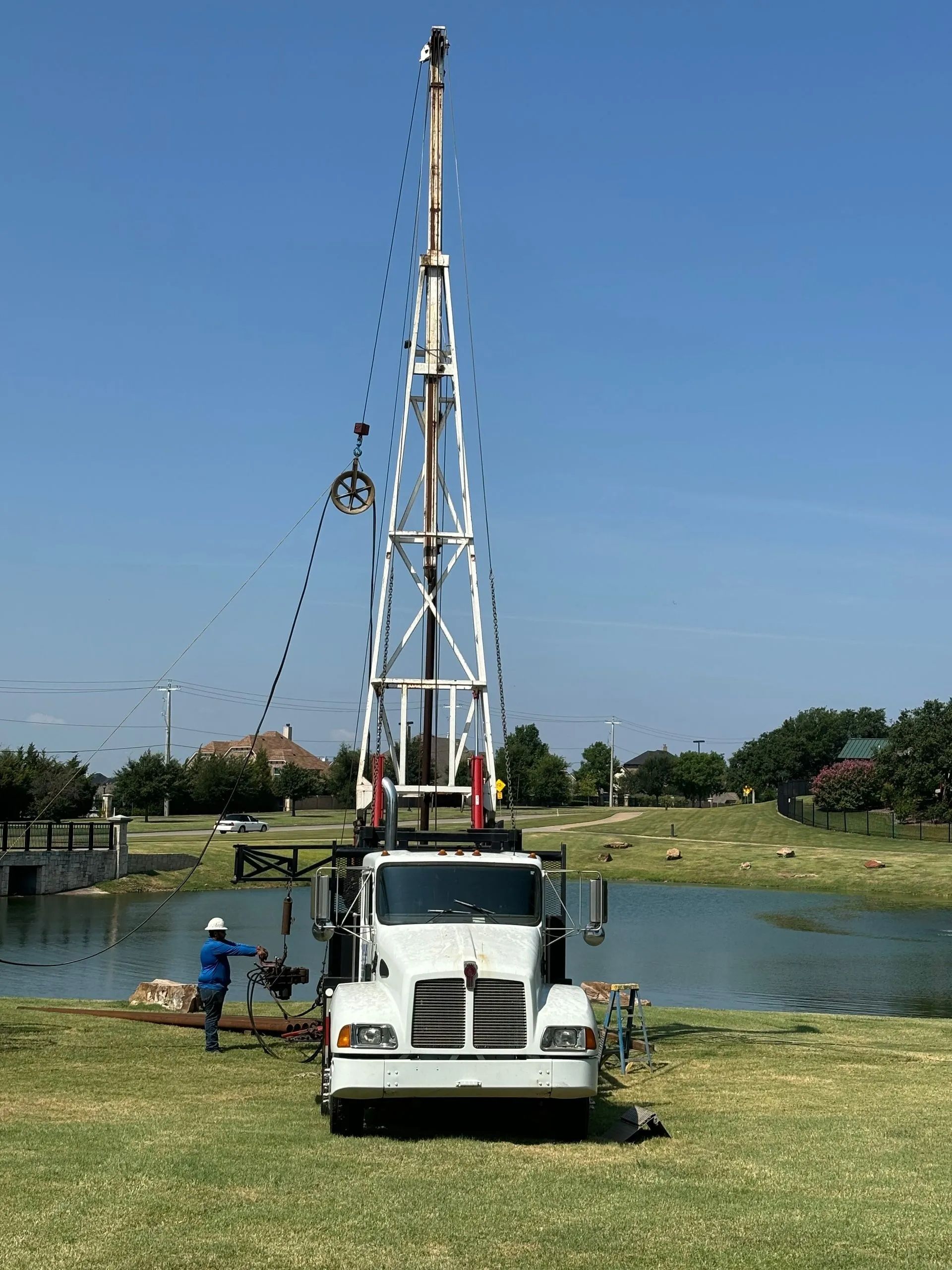 White truck with tall drilling rig beside a pond on a grassy field under a clear blue sky.