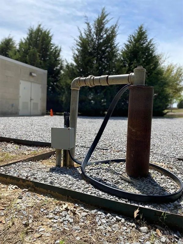 Rusty industrial pipe and valve assembly beside a building, with a black hose on gravel outdoors.