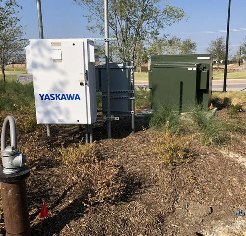 Yaskawa electrical control boxes beside a green utility cabinet outdoors on a dirt embankment