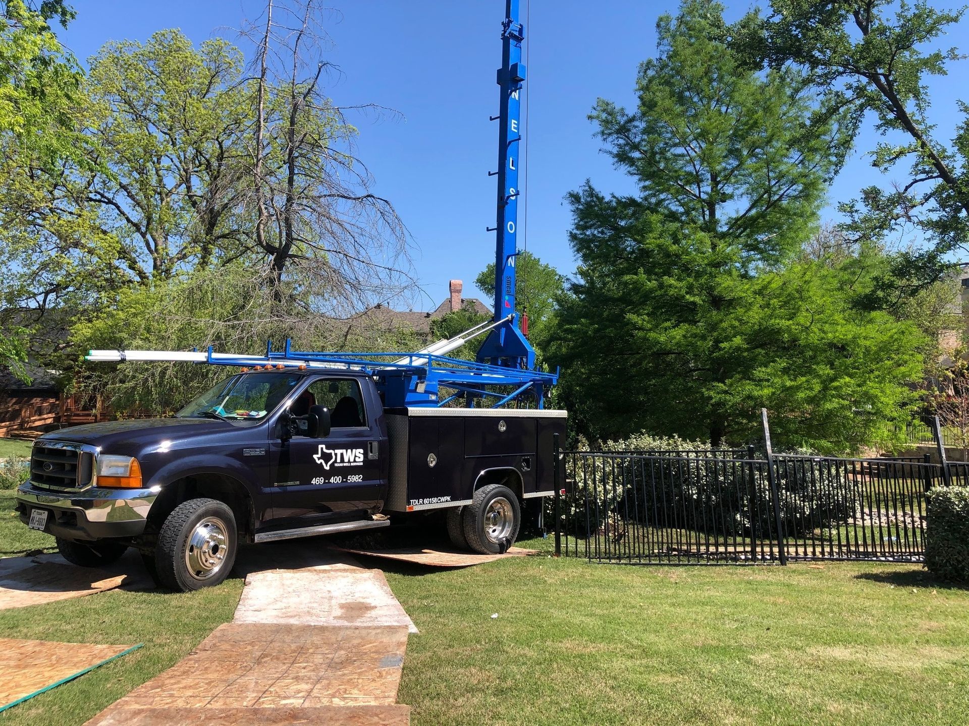 Black utility truck with a blue crane parked on a lawn beside a sidewalk and fence, under trees.