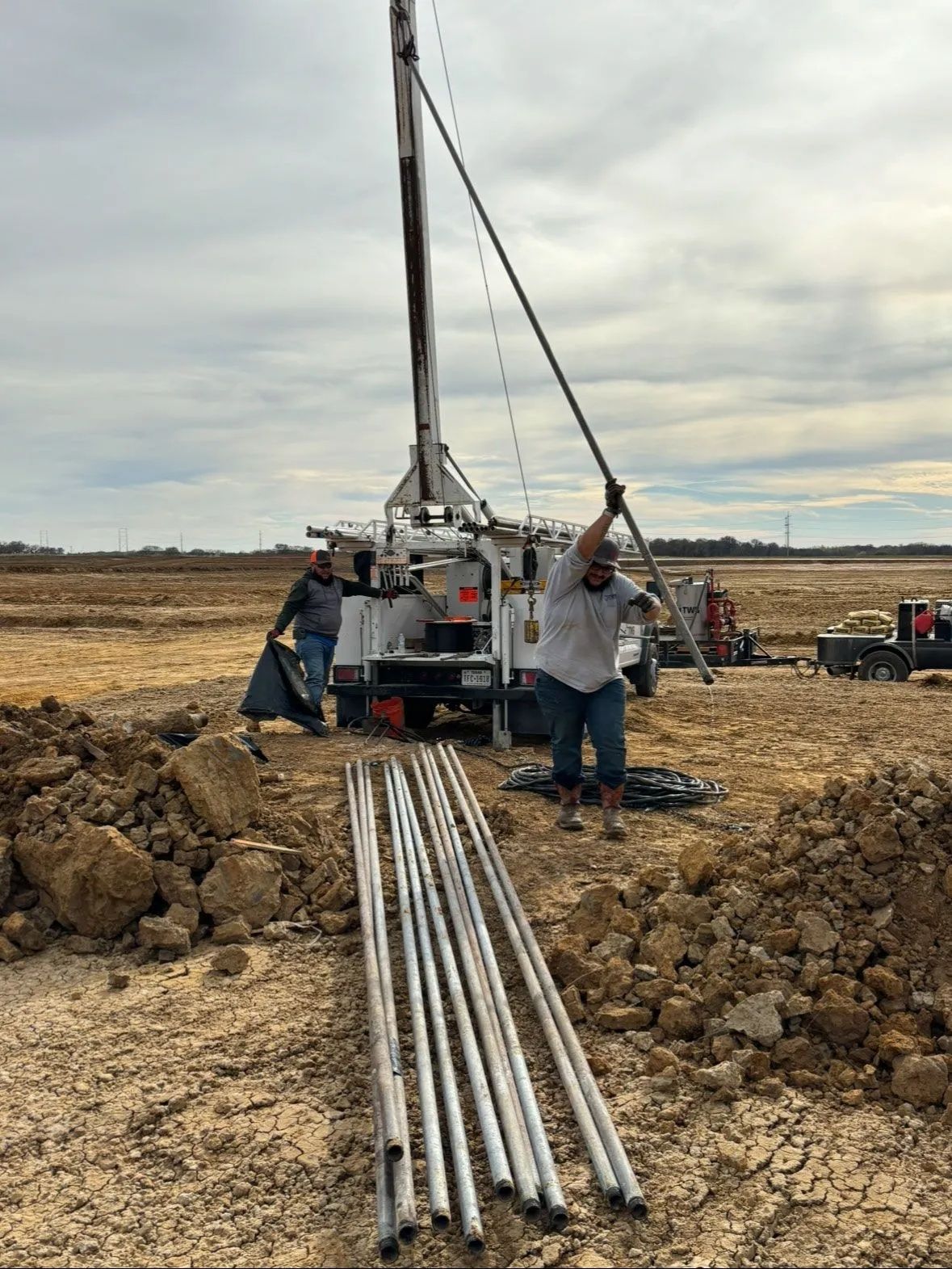 Workers at a drilling rig on a muddy construction site, with pipes laid in the foreground.