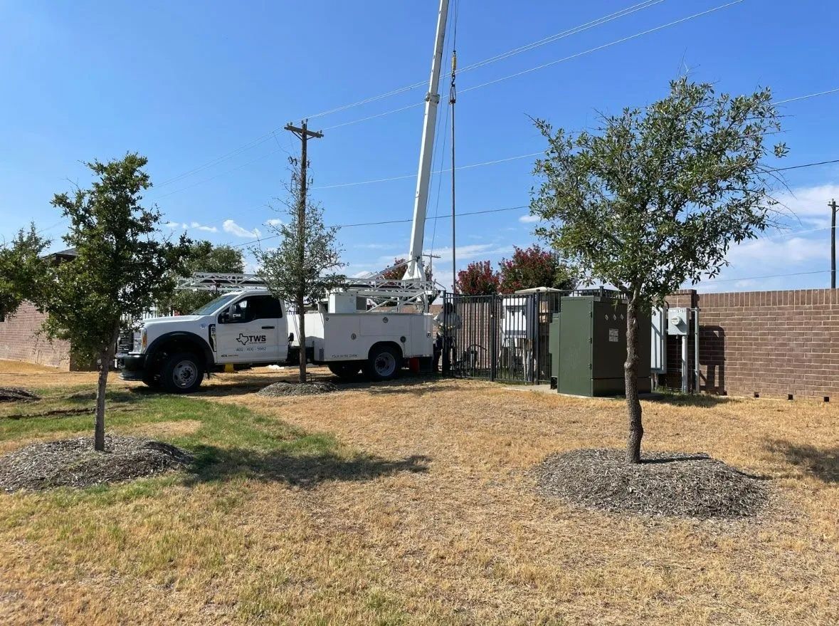 White utility truck and service trailer beside a fenced yard with trees and utility poles under a clear blue sky