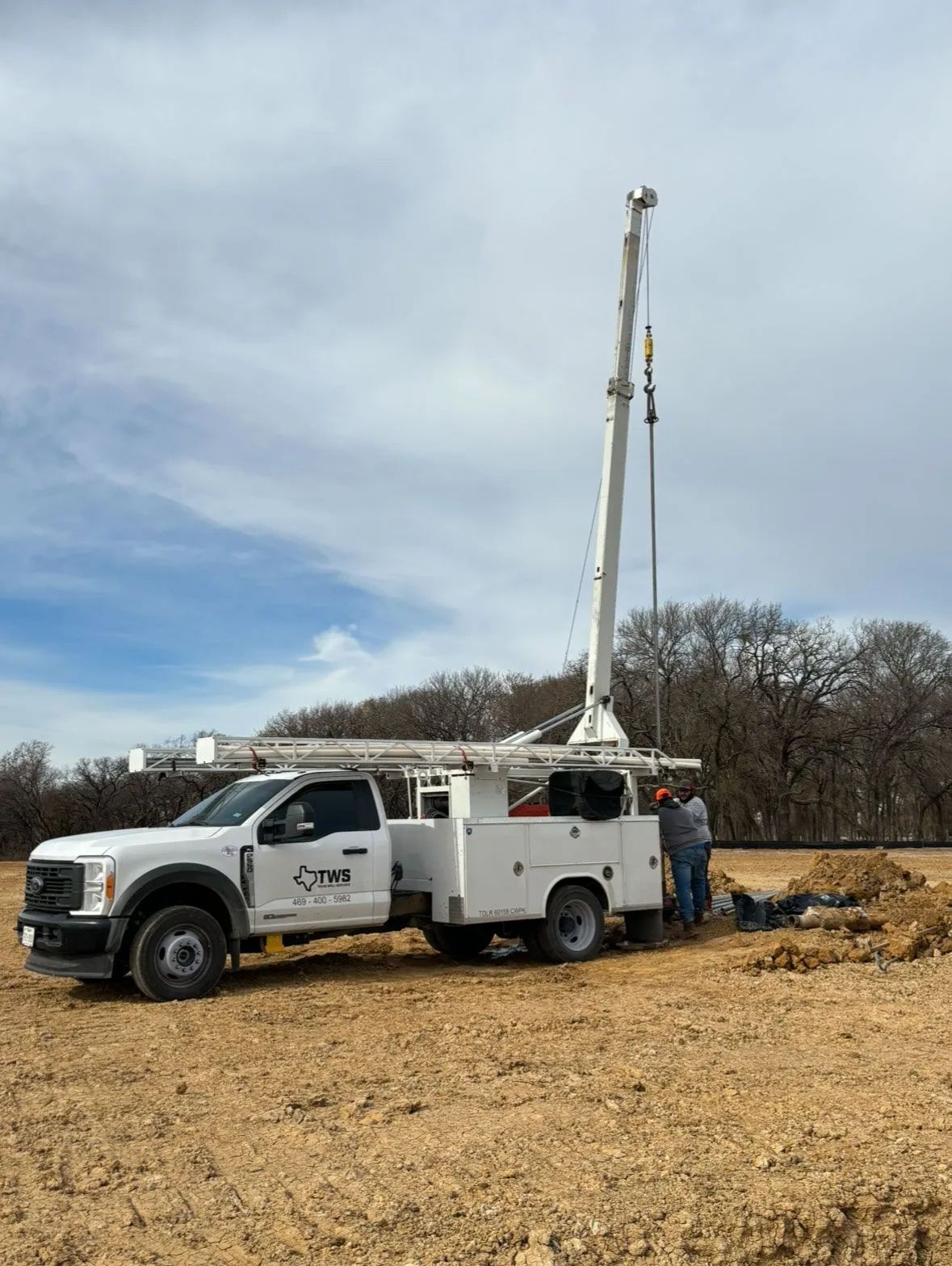 White utility truck with a tall drilling rig parked on a dirt lot under a cloudy sky