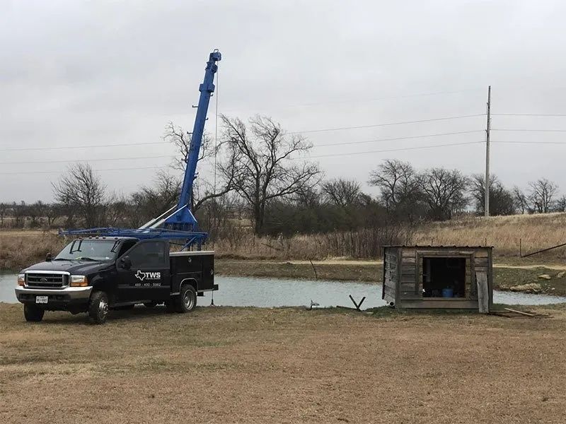 Blue utility truck with raised boom beside a small concrete pit in a grassy field