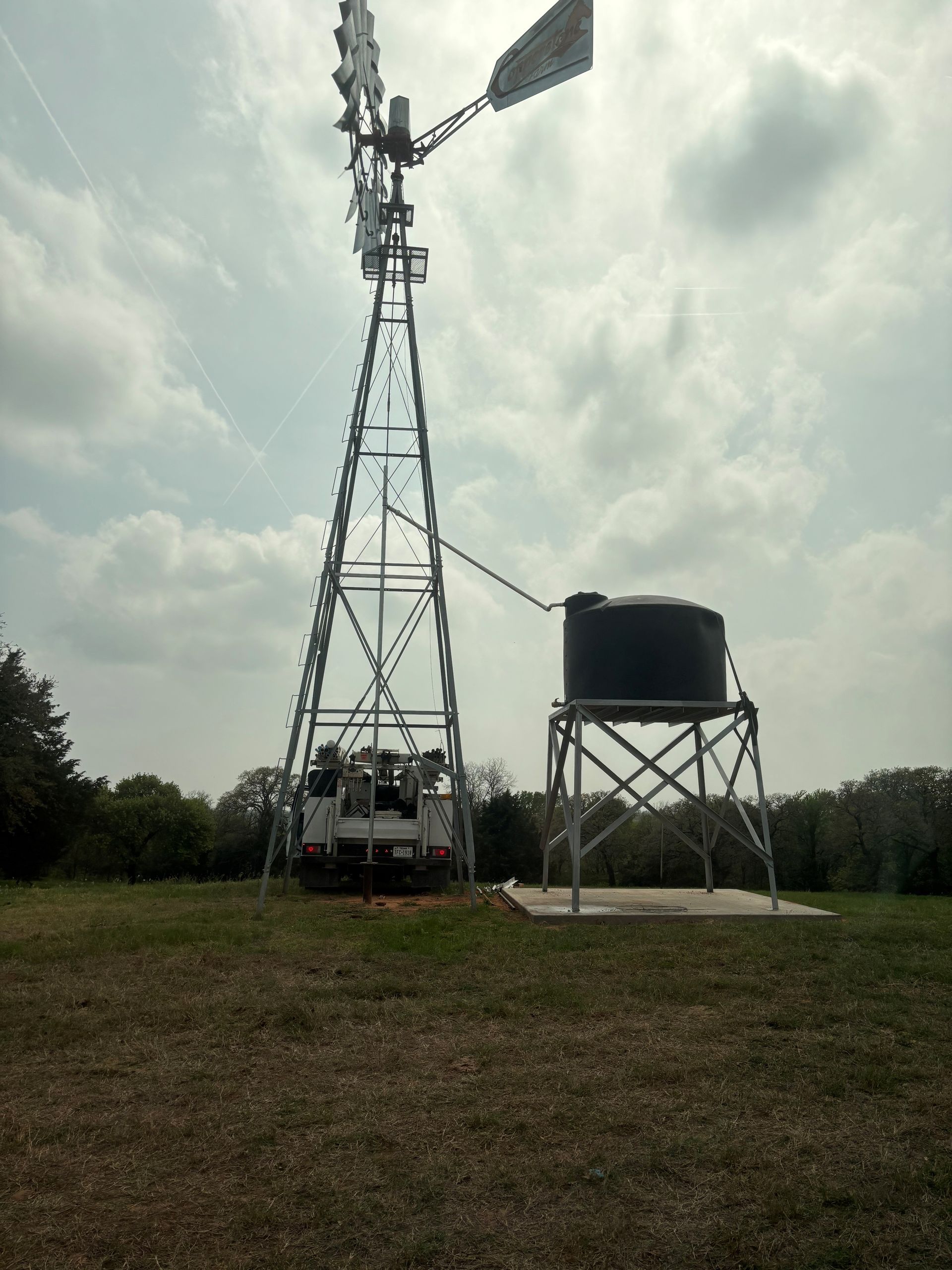 Metal windmill beside a black water tank on a stand in a grassy field under cloudy skies