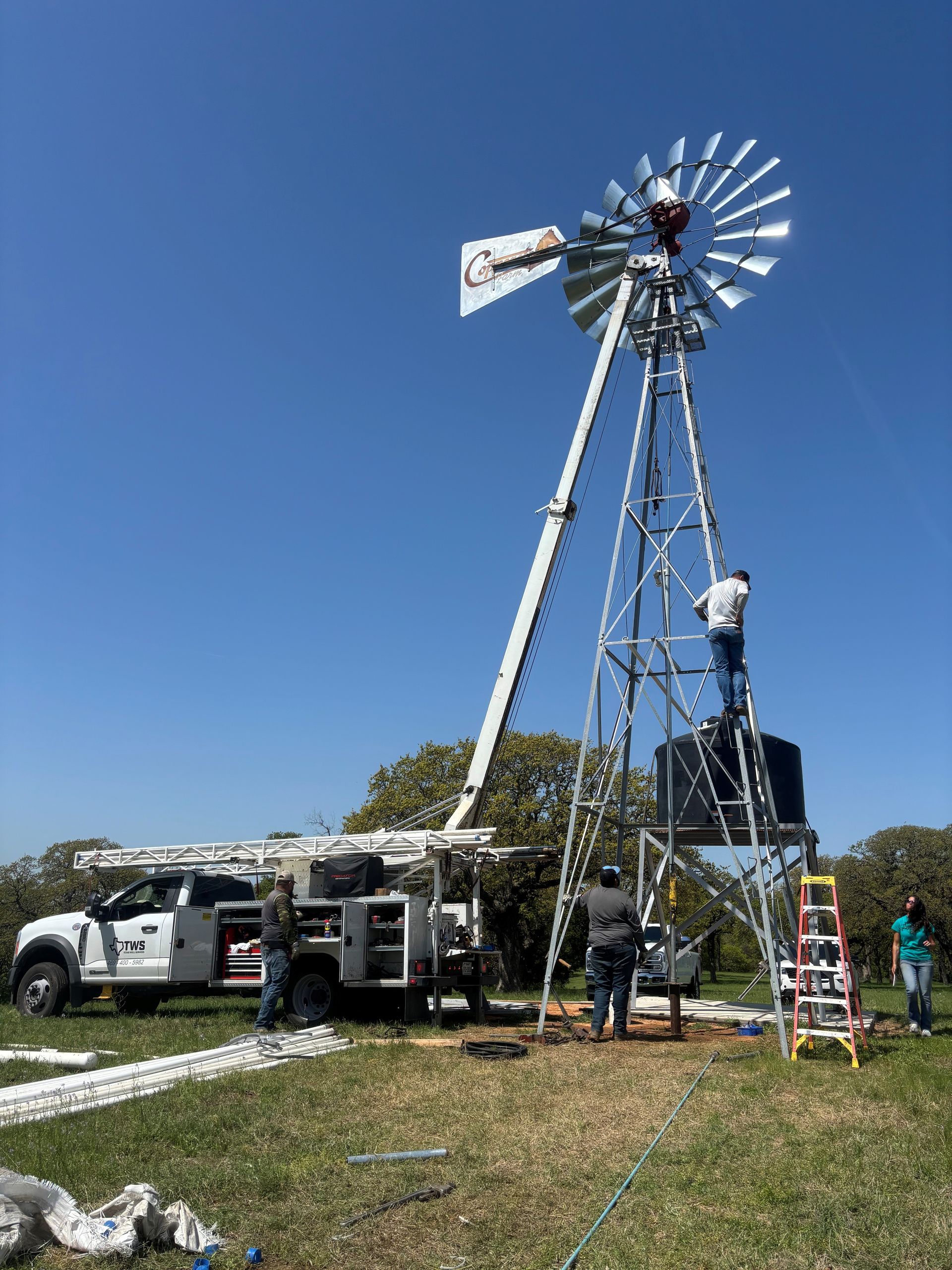 Tall metal windmill tower being repaired outdoors under a clear blue sky, with vehicles and a ladder nearby