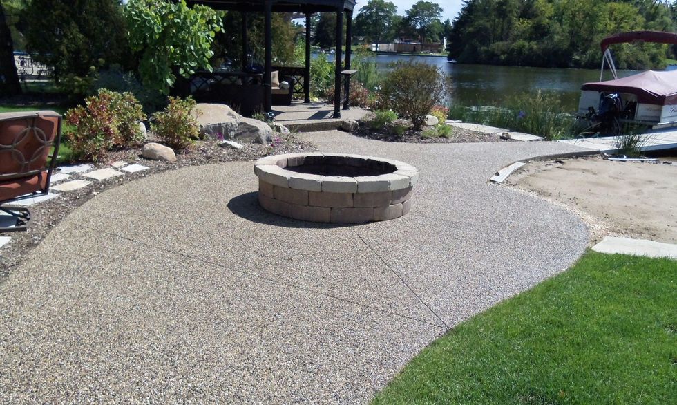 Stone fire pit on a gravel patio beside a river with landscaped plants and green lawn