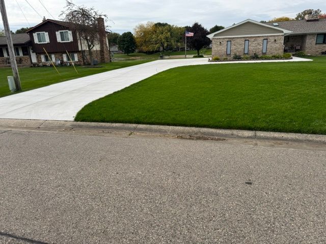 Suburban street with a curved white driveway beside a green lawn and houses on a cloudy day