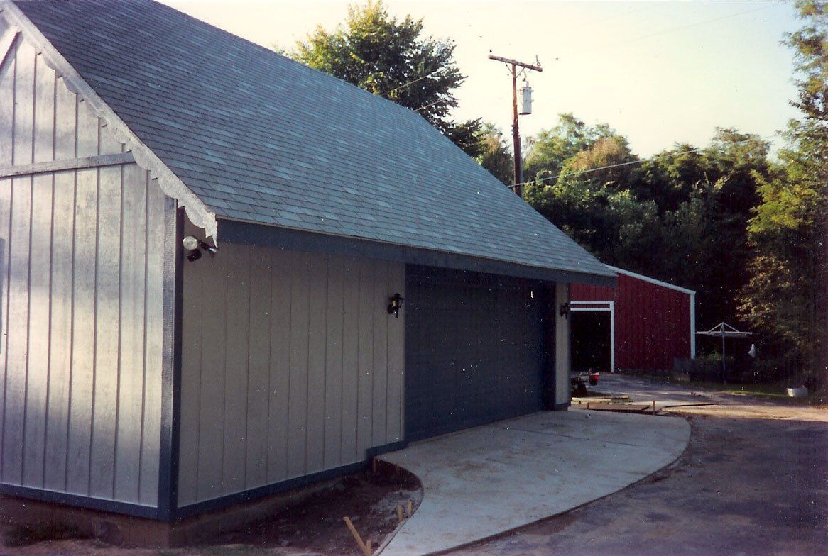 Detached garage with gray siding and dark roof beside a curved driveway, with a red shed in back