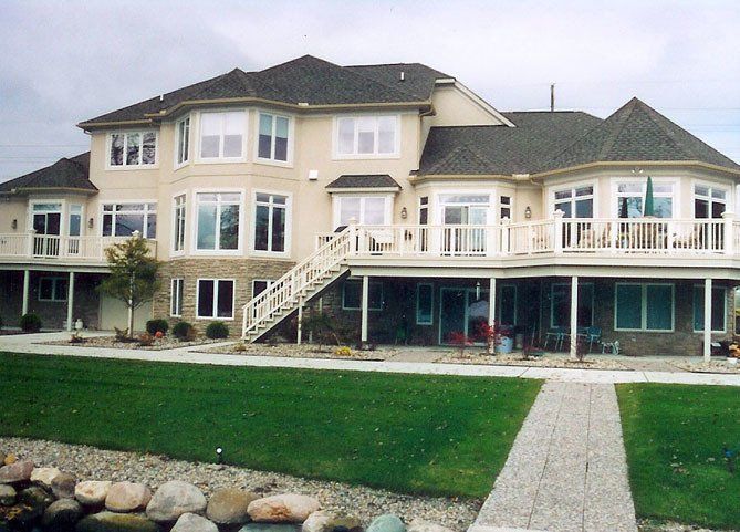 Large beige house with a wraparound porch, white railings, and a green lawn by the water