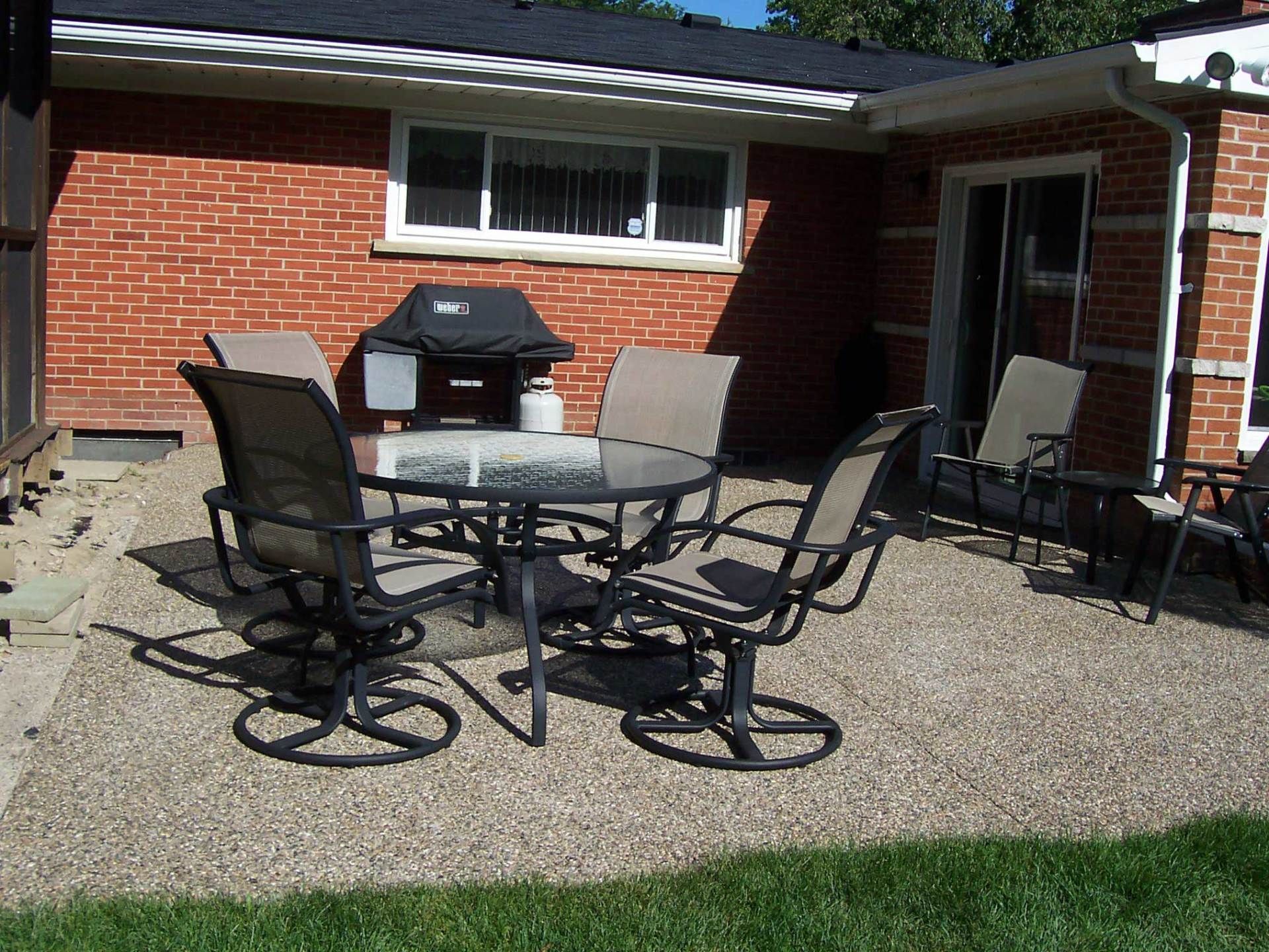 Patio with glass table and chairs on gravel beside a red brick house and grill