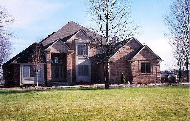Large brick suburban house with a dark roof and leafless trees on a grassy lawn under a clear sky