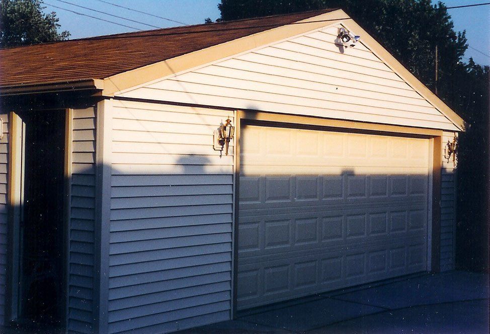 Detached garage with white siding, a tan roll-up door, and a brown shingled roof at sunset
