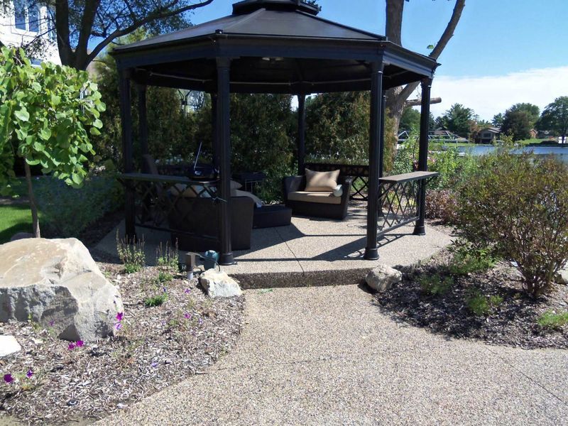 Black gazebo with seating in a landscaped garden beside water under a clear sky