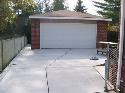 Concrete driveway leading to a closed white garage door, bordered by fences and trees