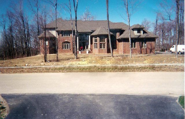 Large brick house with a dark roof and wooded yard, seen across a road on a clear day
