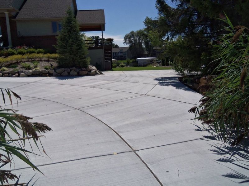 Curved concrete driveway beside a house, with shrubs and trees on a sunny day
