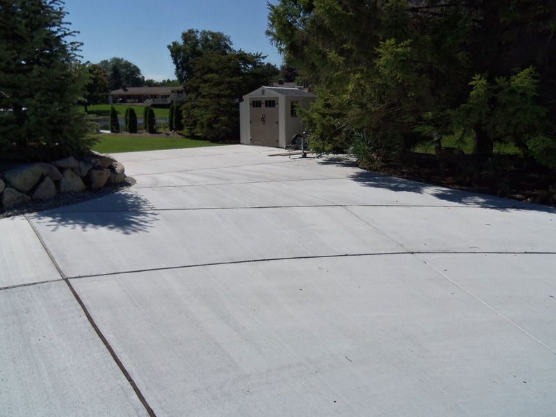 Wide concrete driveway leading to a small building, framed by trees and rocks on a sunny day