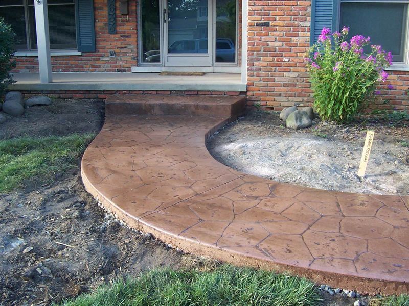 Curved brick walkway under construction in front of a house, with exposed soil and a flower bed on the right.